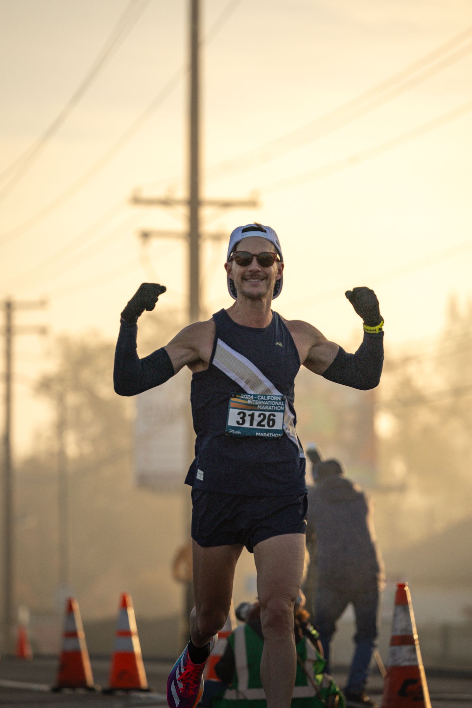 A man in his forties, both arms raised in a bicep flex and grinning, finishing a marathon in hazy amber light.