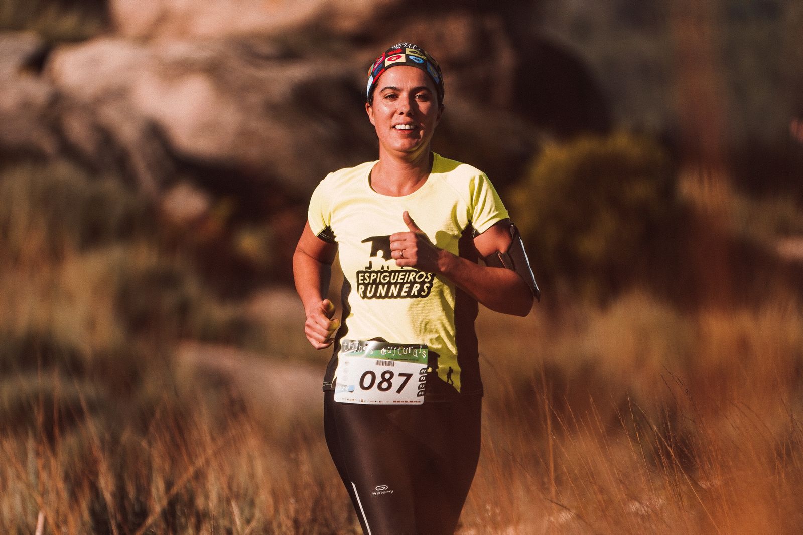 A woman in a club running shirt and headband, race number 087, running through dry golden grass in low evening sun.
