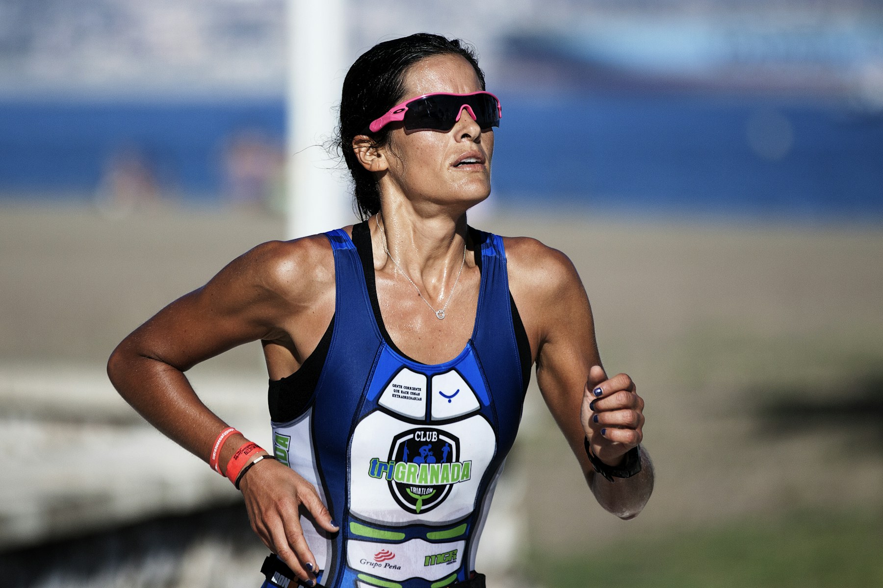 A woman in a club tri-suit and pink sunglasses, sweat on her face and a watch on her wrist, running hard along a seafront.