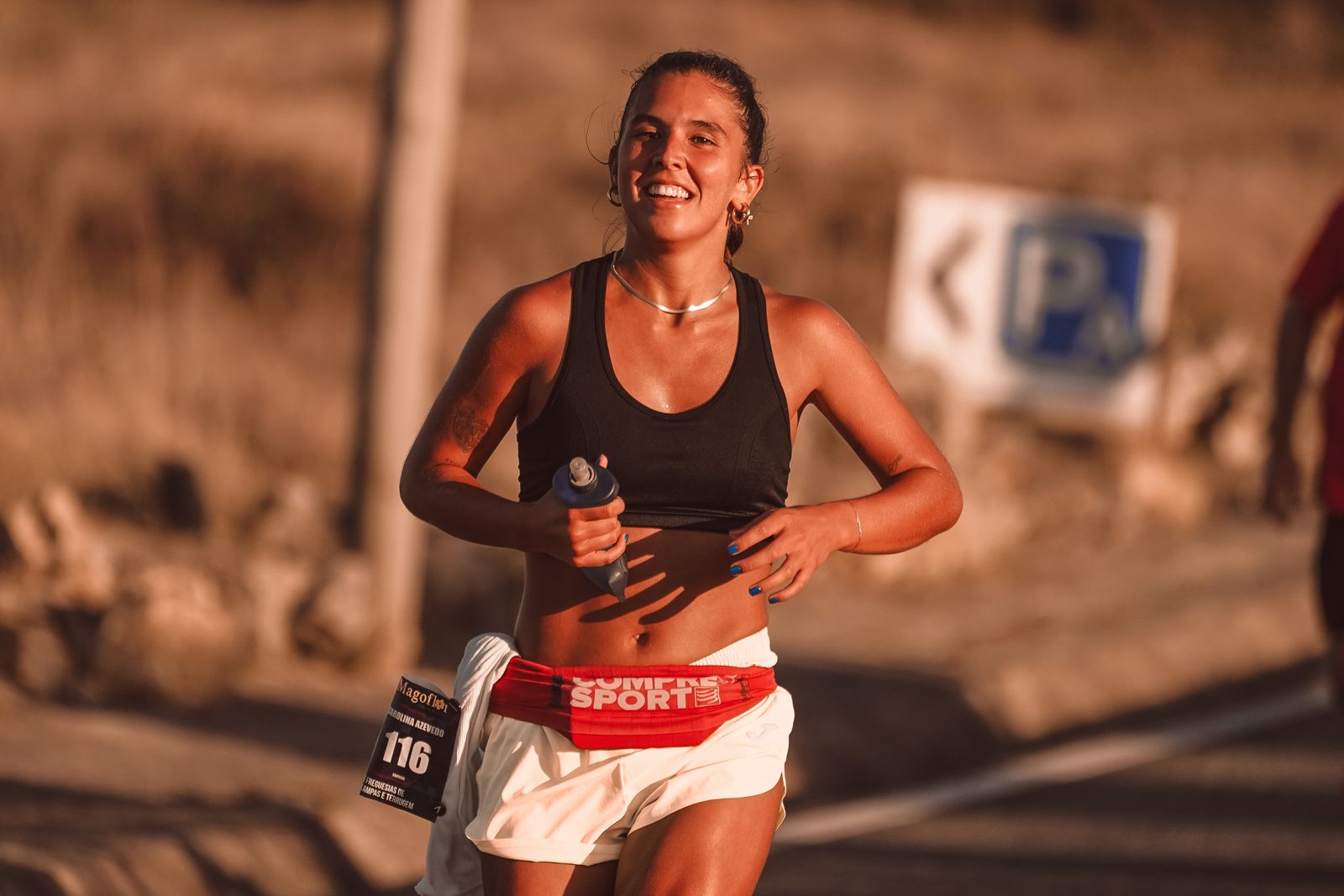 A young woman grinning mid-stride on a road race in warm low sun, a water bottle in one hand.